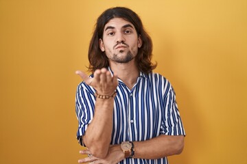 Wall Mural - Hispanic man with long hair standing over yellow background looking at the camera blowing a kiss with hand on air being lovely and sexy. love expression.