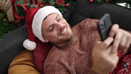 Poster - Young caucasian man using smartphone lying on the sofa by christmas tree at home