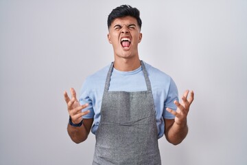 Poster - Hispanic young man wearing apron over white background crazy and mad shouting and yelling with aggressive expression and arms raised. frustration concept.