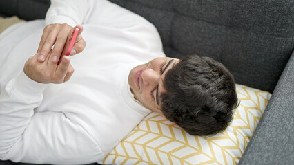 Wall Mural - Young hispanic man using smartphone lying on sofa at home