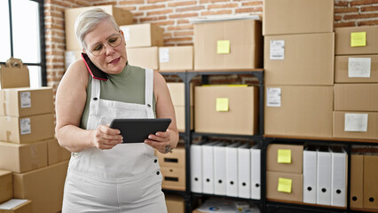 Canvas Print - Middle age grey-haired woman ecommerce business worker talking on smartphone using touchpad at office