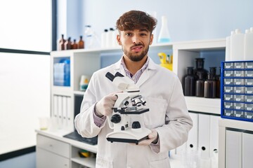 Poster - Arab man with beard working at scientist laboratory holding microscope clueless and confused expression. doubt concept.