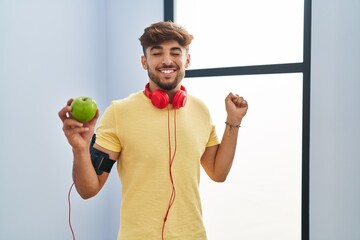 Poster - Arab man with beard wearing sportswear eating green apple screaming proud, celebrating victory and success very excited with raised arm