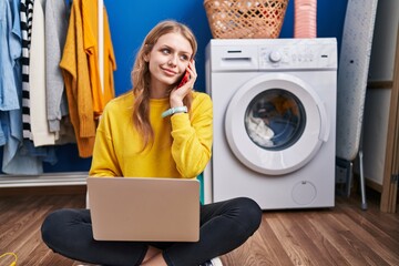 Wall Mural - Young blonde woman using laptop talking on smartphone waiting for washing machine at laundry room