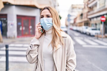 Poster - Young blonde woman wearing medical mask talking on smartphone at street