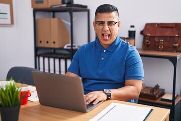 Canvas Print - Young hispanic man working at the office with laptop sticking tongue out happy with funny expression. emotion concept.