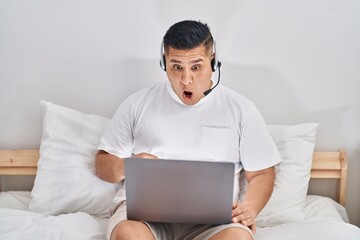 Sticker - Hispanic young man using computer laptop on the bed scared and amazed with open mouth for surprise, disbelief face