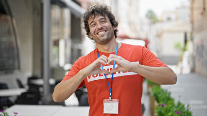 Wall Mural - Young hispanic man activist wearing volunteer uniform doing heart gesture at street