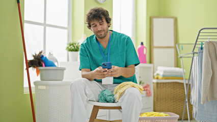 Wall Mural - Young hispanic man professional cleaner using smartphone relaxing at laundry room