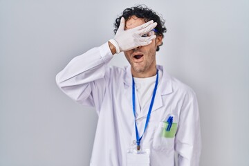 Poster - Hispanic man working at scientist laboratory peeking in shock covering face and eyes with hand, looking through fingers afraid