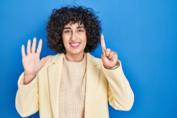 Wall Mural - Young brunette woman with curly hair standing over blue background showing and pointing up with fingers number six while smiling confident and happy.