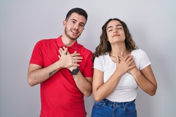 Canvas Print - Young hispanic couple standing over isolated background smiling with hands on chest with closed eyes and grateful gesture on face. health concept.