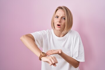 Wall Mural - Young caucasian woman standing over pink background in hurry pointing to watch time, impatience, upset and angry for deadline delay