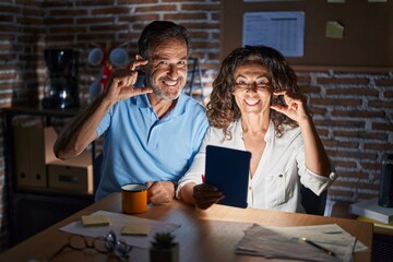 Canvas Print - Middle age hispanic couple using touchpad sitting on the table at night smiling and confident gesturing with hand doing small size sign with fingers looking and the camera. measure concept.