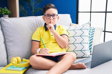 Poster - Young hispanic kid doing homework sitting on the sofa asking to be quiet with finger on lips. silence and secret concept.