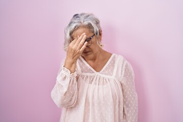 Middle age woman with grey hair standing over pink background tired rubbing nose and eyes feeling fatigue and headache. stress and frustration concept.