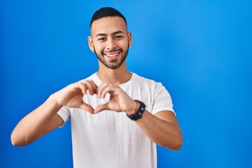 Canvas Print - Young hispanic man standing over blue background smiling in love doing heart symbol shape with hands. romantic concept.