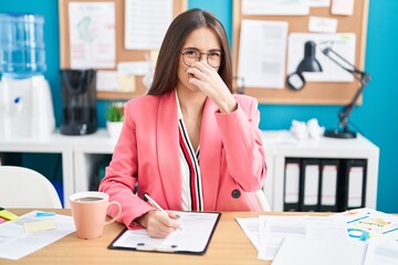 Sticker - Young hispanic woman working at the office wearing glasses smelling something stinky and disgusting, intolerable smell, holding breath with fingers on nose. bad smell