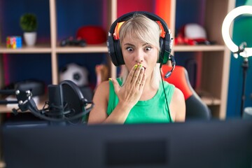 Poster - Young caucasian woman playing video games wearing headphones covering mouth with hand, shocked and afraid for mistake. surprised expression