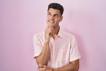 Poster - Young hispanic man standing over pink background with hand on chin thinking about question, pensive expression. smiling with thoughtful face. doubt concept.