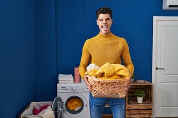 Wall Mural - Young hispanic man holding laundry basket sticking tongue out happy with funny expression.