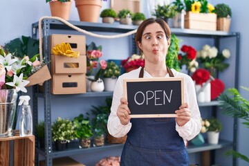 Sticker - Brunette woman working at florist holding open sign making fish face with mouth and squinting eyes, crazy and comical.