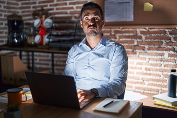 Canvas Print - Hispanic man with beard working at the office at night puffing cheeks with funny face. mouth inflated with air, crazy expression.