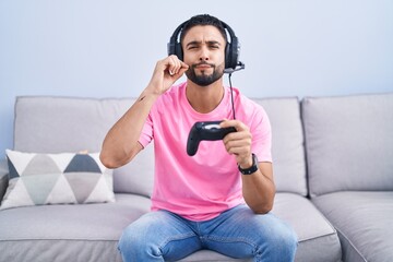 Canvas Print - Hispanic young man playing video game holding controller sitting on the sofa mouth and lips shut as zip with fingers. secret and silent, taboo talking