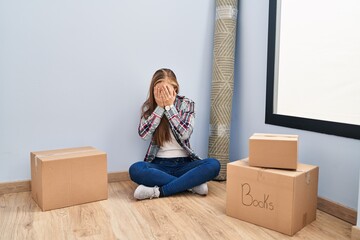 Poster - Young blonde woman sitting on the floor moving to a new home with sad expression covering face with hands while crying. depression concept.