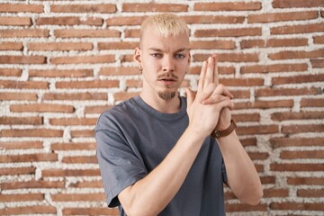 Poster - Young caucasian man standing over bricks wall holding symbolic gun with hand gesture, playing killing shooting weapons, angry face