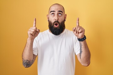 Canvas Print - Young hispanic man with beard and tattoos standing over yellow background amazed and surprised looking up and pointing with fingers and raised arms.