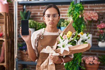 Wall Mural - Young hispanic woman working at florist shop showing smartphone screen skeptic and nervous, frowning upset because of problem. negative person.
