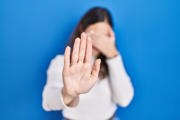 Sticker - Young hispanic woman standing over blue background covering eyes with hands and doing stop gesture with sad and fear expression. embarrassed and negative concept.