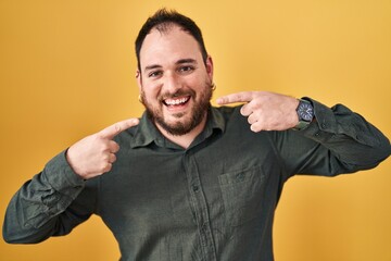 Wall Mural - Plus size hispanic man with beard standing over yellow background smiling cheerful showing and pointing with fingers teeth and mouth. dental health concept.