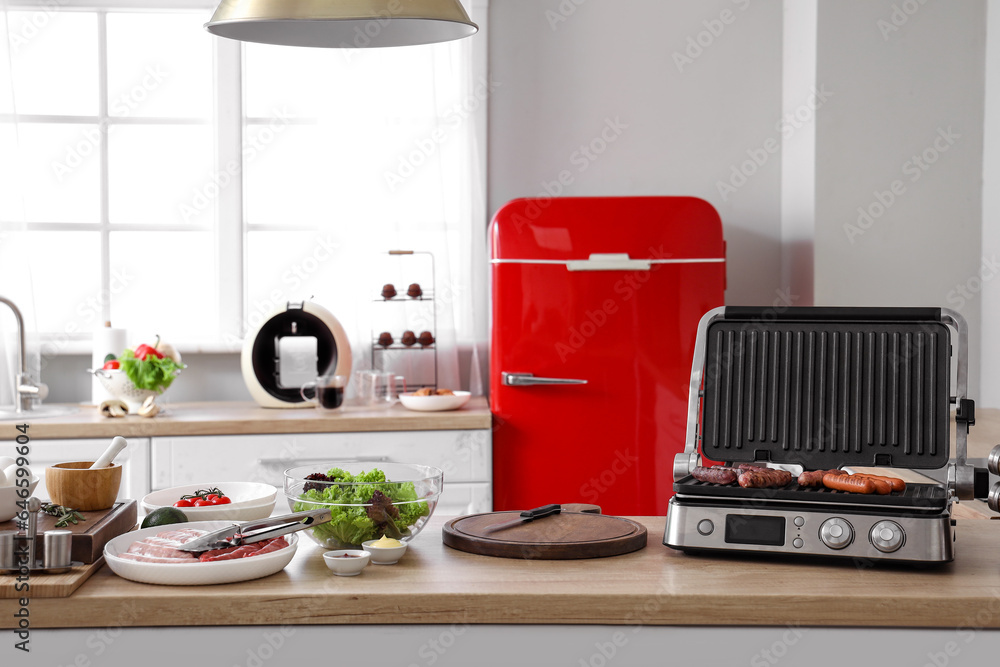 Electric grill with tasty sausages on table in kitchen