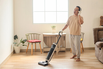 Wall Mural - Happy young man wearing wireless headphones cleaning home with vacuum cleaner.