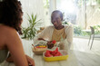 © DragonImages - Smiling Black young woman eating breakfast in kitchen of her friend
