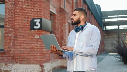 Arabian man with serious facial expression standing on fresh air and working on remote computer. Pensive young male wearing comfy sky blue shirt thoughtfully looking up and then typing on keyboard.