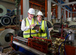 © chadchai - Two worker standing amidst the humming machinery of a bustling factory. Discover the essence of teamwork, innovation and precision as these skilled individuals navigate the industrial landscape.