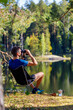 A man with binoculars on the lake shore in the autumn forest early in the morning, the background is blurred. A coffee maker with a cup of coffee is nearby.