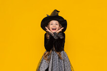 A Funny And Joyful Child In A Halloween Costume, On An Isolated Yellow Background. A Happy Little Girl In A Witch Costume And A Witch Hat Is Having Fun At A Halloween Party.