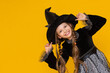 © Юлия Дьякова - A young witch in a black fluffy dress and long pigtails supports her pointed black hat with her hands. portrait of an attractive witch on a yellow isolated background.