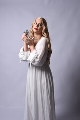 Wall Mural - Close up portrait of beautiful blonde model wearing elegant  white halloween gown, a historical fantasy character.  Holding crucifix cross necklace, isolated on studio background.