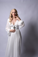 Wall Mural - Close up portrait of beautiful blonde model wearing elegant  white halloween gown, a historical fantasy character.  Holding crucifix cross necklace, isolated on studio background.