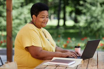 Side view portrait of modern Black senior woman using laptop at outdoor cafe and working, copy space