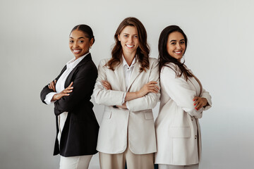 Successful international young businesswomen posing with arms crossed and smiling at camera, standing over light wall