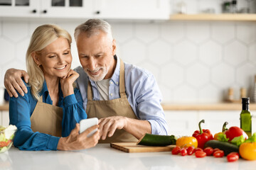 Smiling senior spouses using smartphone in kitchen while cooking food together