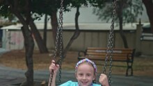 Joyful Little Girl Rides Swings Holding By Chains In City Park Smiling Child With Headband Plays Swinging Attraction On Playground Schoolgirl Swings In Yard