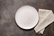 Empty white plate with a beige linen napkin on a brown rustic background. Top view, flat lay, copy space.