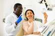 Positive asian woman and african-american man dentist looking at teeth through mirror after dental restoration procedure.